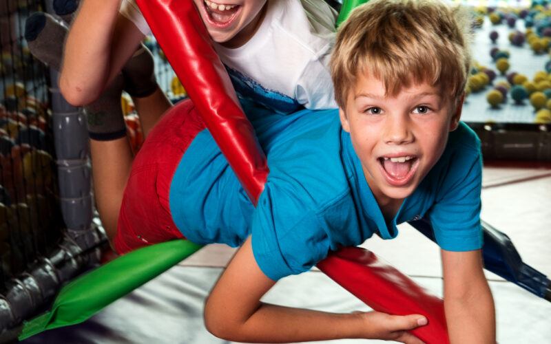 Zwei kleine Jungen spielen und lächeln, während sie auf bunten gepolsterten Stangen in einem Indoor-Spielbereich klettern.