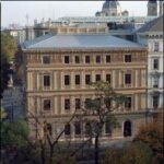 Ein dreistöckiges historisches Steingebäude mit Bogenfenstern und kunstvollen architektonischen Details, umgeben von Bäumen und benachbarten Gebäuden.