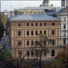 Ein dreistöckiges historisches Steingebäude mit Bogenfenstern und kunstvollen architektonischen Details, umgeben von Bäumen und benachbarten Gebäuden.