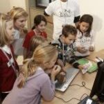 Eine Gruppe von Kindern versammelt sich unter Aufsicht eines Erwachsenen um einen Computermonitor, während sie an einem Schreibtisch eine Tätigkeit ausüben.