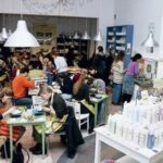 Ein belebtes Café mit Menschen, die an Tischen sitzen, essen und sich unterhalten; Regale mit Produkten sind sichtbar, und helle Oberlichter erhellen den Raum.