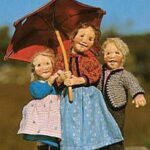 Drei Puppen stehen im Freien dicht beieinander, wobei die mittlere Puppe einen roten Regenschirm über alle hält.