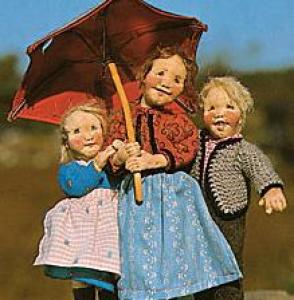 Drei Puppen stehen im Freien dicht beieinander, wobei die mittlere Puppe einen roten Regenschirm über alle hält.