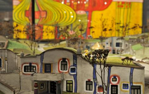 Modell eines farbenfrohen, abstrakten Gebäudes mit geschwungenen Linien und quadratischen Fenstern, eingebettet in eine Miniaturlandschaft mit gemalten Bäumen und einer lebendigen, künstlerischen Kulisse.