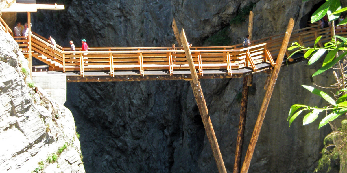 Eine hölzerne Fußgängerbrücke mit abgewinkelten Stützbalken überspannt eine felsige Schlucht über einem fließenden Fluss, über die Menschen gehen.