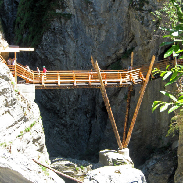 Eine hölzerne Fußgängerbrücke mit abgewinkelten Stützbalken überspannt eine felsige Schlucht über einem fließenden Fluss, über die Menschen gehen.