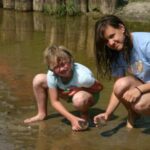 Zwei Mädchen kauern am Rande eines seichten, schlammigen Baches, untersuchen Gegenstände in ihren Händen und schauen in die Kamera.