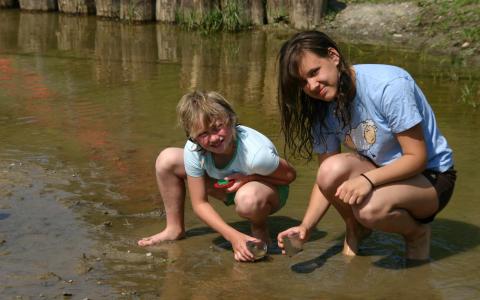 Zwei Mädchen kauern am Rande eines seichten, schlammigen Baches, untersuchen Gegenstände in ihren Händen und schauen in die Kamera.