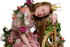 Eine junge Frau mit Krone und rosafarbenem, blumengeschmücktem Kleid schläft, den Kopf auf ein Spinnrad gestützt.