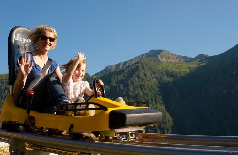 Zwei Personen, ein Erwachsener und ein Kind, fahren lächelnd und winkend mit Bergen und Bäumen im Hintergrund auf einer gelben Alpen-Achterbahn im Freien.