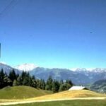 Eine Seilbahn fährt über einen grasbewachsenen Hang mit vereinzelten Bäumen, Almhütten und entfernten schneebedeckten Bergen unter einem klaren blauen Himmel.