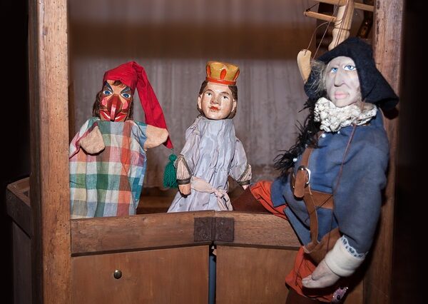 Drei Handpuppen sind auf einer kleinen hölzernen Puppentheaterbühne angeordnet, darunter eine Figur mit rotem Hut, eine Königin mit Krone und eine Figur in blauem Gewand mit Federhut.