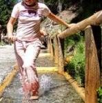 Eine Person läuft barfuß durch einen seichten Wasserlauf auf einem Steinweg und hält sich dabei an einem Holzgeländer im Freien fest.