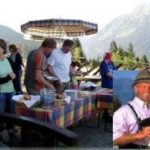 An einem Buffet im Freien in den Bergen werden Speisen serviert. Eine Einblendung zeigt einen Mann in traditioneller Alpenkleidung, der eine Holzflöte spielt.