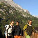 Drei Personen in Wanderkleidung gehen an einem sonnigen Tag im Freien spazieren, mit Bergen und Bäumen im Hintergrund.