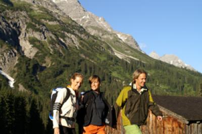 Drei Personen in Wanderkleidung gehen an einem sonnigen Tag im Freien spazieren, mit Bergen und Bäumen im Hintergrund.