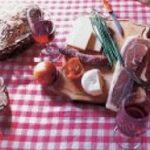 Auf einem Holzbrett auf einer rot karierten Tischdecke liegen Brot, Käse, Wurst, grüne Zwiebeln, Tomaten und ein Apfel, daneben zwei Gläser Rotwein.