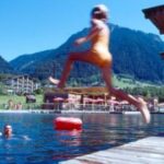 Ein Kind im Badeanzug springt von einem Steg in einen See, während eine Person vom Wasser aus winkt, mit Bergen und Gebäuden im Hintergrund.