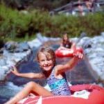 Ein Kind in einem Badeanzug sitzt auf einem roten Schlauch und schwimmt einen kleinen künstlichen Wasserkanal hinunter, während ein anderes Kind im Hintergrund zu sehen ist.
