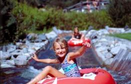Ein Kind in einem Badeanzug sitzt auf einem roten Schlauch und schwimmt einen kleinen künstlichen Wasserkanal hinunter, während ein anderes Kind im Hintergrund zu sehen ist.