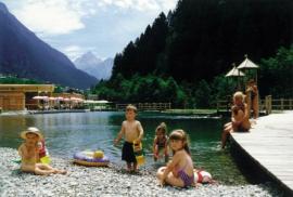 Kinder spielen am Rande eines Sees, der von Bergen umgeben ist, während die Erwachsenen auf einer Holzterrasse in der Nähe unter einem sonnigen Himmel sitzen.