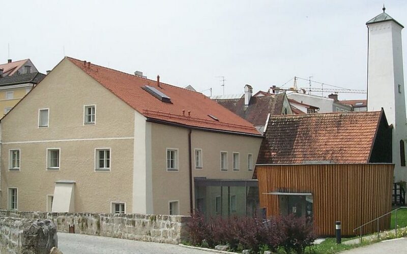 Eine Gruppe von Gebäuden mit roten Ziegeldächern, darunter ein beigefarbenes verputztes Haus und ein holzgetäfeltes Gebäude neben einem hohen, weißen rechteckigen Turm.