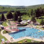 Freibad mit Menschen, die schwimmen und sich sonnen, umgeben von Rasenflächen, Bäumen und Hügeln im Hintergrund an einem sonnigen Tag.