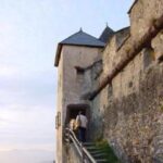 Eine Person geht bei teilweise bewölktem Himmel über steinerne Stufen auf den Eingang eines alten Schlosses mit hohen Steinmauern zu.