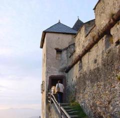 Eine Person geht bei teilweise bewölktem Himmel über steinerne Stufen auf den Eingang eines alten Schlosses mit hohen Steinmauern zu.