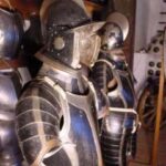 Zwei mittelalterliche Rüstungen mit Helmen und Visieren sind in einem Museum nebeneinander ausgestellt, umgeben von anderen historischen Rüstungsteilen.