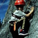 Ein Kind, das einen Helm und einen Klettergurt trägt, klettert im Freien an einer Felswand, hält sich an Griffen fest und trägt Sportkleidung.