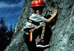Ein Kind, das einen Helm und einen Klettergurt trägt, klettert im Freien an einer Felswand, hält sich an Griffen fest und trägt Sportkleidung.