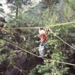Zwei Personen überqueren eine Schlucht in einem Wald mit Hilfe einer Seilbrücke, wobei sie sich an den oberen Seilen festhalten, um sich abzustützen und das Gleichgewicht zu halten.