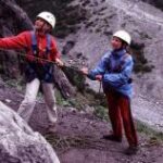 Zwei Personen mit Helm und Klettergurt stehen in felsigem Gelände; einer steigt die Felswand hinauf, während der andere das Seil zur Sicherung hält.