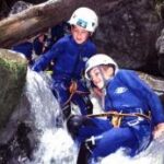 Zwei Personen, die Helme und Neoprenanzüge tragen, navigieren in einem felsigen Bach, umgeben von Wasser und großen Felsbrocken in einer Außenumgebung.