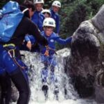 Eine Gruppe von Personen in Neoprenanzügen und Helmen steht beim Canyoning im flachen Wasser zwischen Felsen und wird dabei von einem Führer unterstützt.