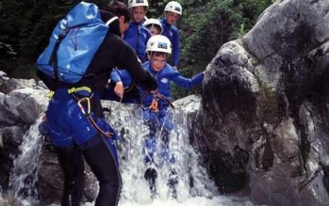 Eine Gruppe von Personen in Neoprenanzügen und Helmen steht beim Canyoning im flachen Wasser zwischen Felsen und wird dabei von einem Führer unterstützt.