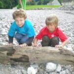 Zwei kleine Kinder kauern auf einer felsigen Fläche im Freien und stützen sich jeweils mit einer Hand auf ein großes Stück Treibholz.