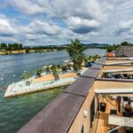 Eine Szene am Flussufer mit einem Steg, Sitzgelegenheiten und Menschen, die sich am Wasser unter einem teilweise bewölkten Himmel entspannen. Gebäude und Bäume säumen das gegenüberliegende Flussufer.