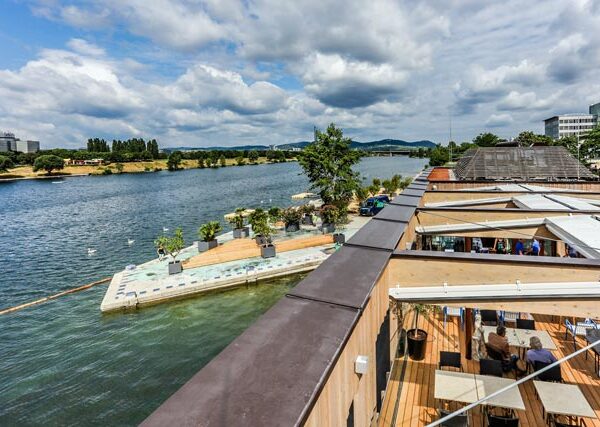 Eine Szene am Flussufer mit einem Steg, Sitzgelegenheiten und Menschen, die sich am Wasser unter einem teilweise bewölkten Himmel entspannen. Gebäude und Bäume säumen das gegenüberliegende Flussufer.