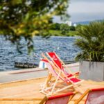 Mehrere rote Liegestühle und eine Topfpalme stehen auf einer Sandfläche am Wasser, während im Hintergrund Gebäude und Grünflächen zu sehen sind.