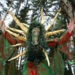 Person in einem Wald, die ein grün-rotes Kostüm mit goldenen Stacheln auf dem Rücken trägt, die Arme erhoben und das Gesicht grün bemalt.