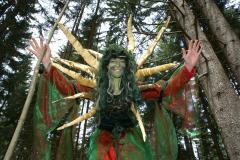 Person in einem Wald, die ein grün-rotes Kostüm mit goldenen Stacheln auf dem Rücken trägt, die Arme erhoben und das Gesicht grün bemalt.