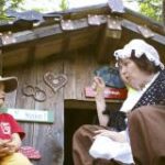 Eine ältere Frau in einem Kostüm spricht mit einem Kind, das vor einem kleinen Holzhaus sitzt, das mit Herz- und Brillenschildern geschmückt ist.