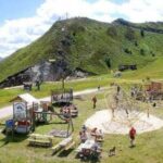 Kinder und Erwachsene erfreuen sich an einem Spielplatz mit Klettergerüsten und Picknicktischen auf einer grasbewachsenen Anhöhe, mit Bergen im Hintergrund unter einem teilweise bewölkten Himmel.