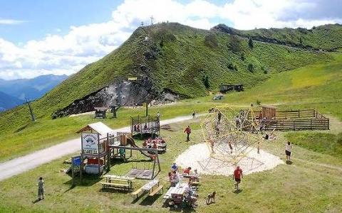 Kinder und Erwachsene erfreuen sich an einem Spielplatz mit Klettergerüsten und Picknicktischen auf einer grasbewachsenen Anhöhe, mit Bergen im Hintergrund unter einem teilweise bewölkten Himmel.