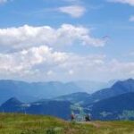 Menschen stehen auf einer grasbewachsenen Anhöhe und blicken unter einem teilweise bewölkten Himmel auf ferne blaue Berge.