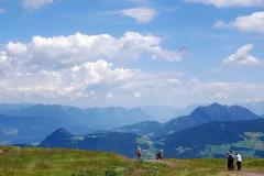Menschen stehen auf einer grasbewachsenen Anhöhe und blicken unter einem teilweise bewölkten Himmel auf ferne blaue Berge.