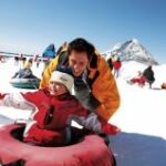 Ein Erwachsener und ein Kind in Winterkleidung genießen Snowtubing auf einem verschneiten Hügel mit anderen Menschen und einem Berg im Hintergrund.