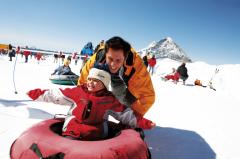 Ein Erwachsener und ein Kind in Winterkleidung genießen Snowtubing auf einem verschneiten Hügel mit anderen Menschen und einem Berg im Hintergrund.